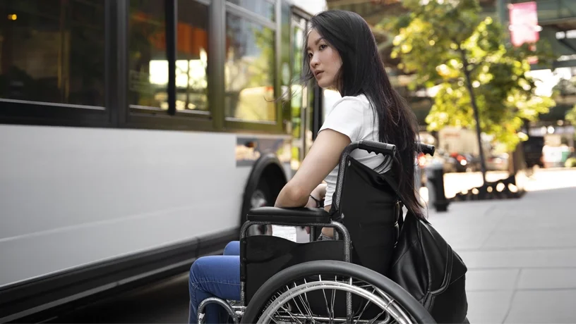 Junge Frau mit ALS im Rollstuhl an einer Busstation