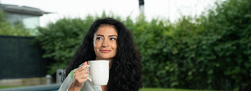 Frau mit lockigen Haaren sitzt vor einer grünen Hecke und hält eine Teetasse und schaut selbstbewußt.