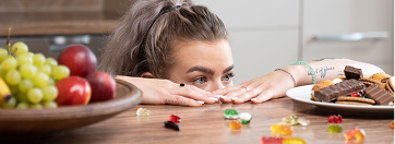 Frau schaut über Tischkante auf Obst, Süßigkeiten und herzhafte Snacks